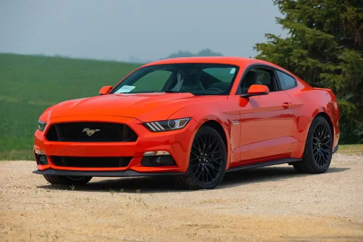 2015 Ford Mustang GT for Sale at Auction - Mecum Auctions - Engine Stories Bright orange sports car parked on a rural dirt road against a backdrop of green fields and trees.