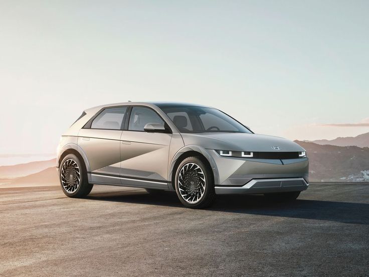 Sleek silver electric car parked on a scenic road with mountains in the background under a clear sky.