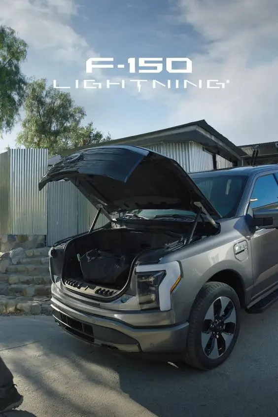 90321eb1231624e1e2938d49a1ff474f - Engine Stories Ford F-150 Lightning with open hood showcasing storage space, parked outdoors on a sunny day. Electric truck front view.