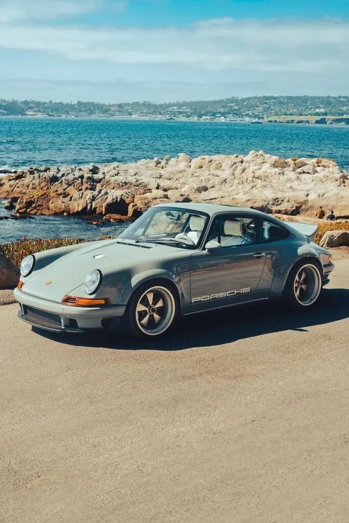 DLS - Engine Stories Vintage Porsche parked by a rocky coastal landscape with ocean view under a clear blue sky. Classic car, scenic drive.