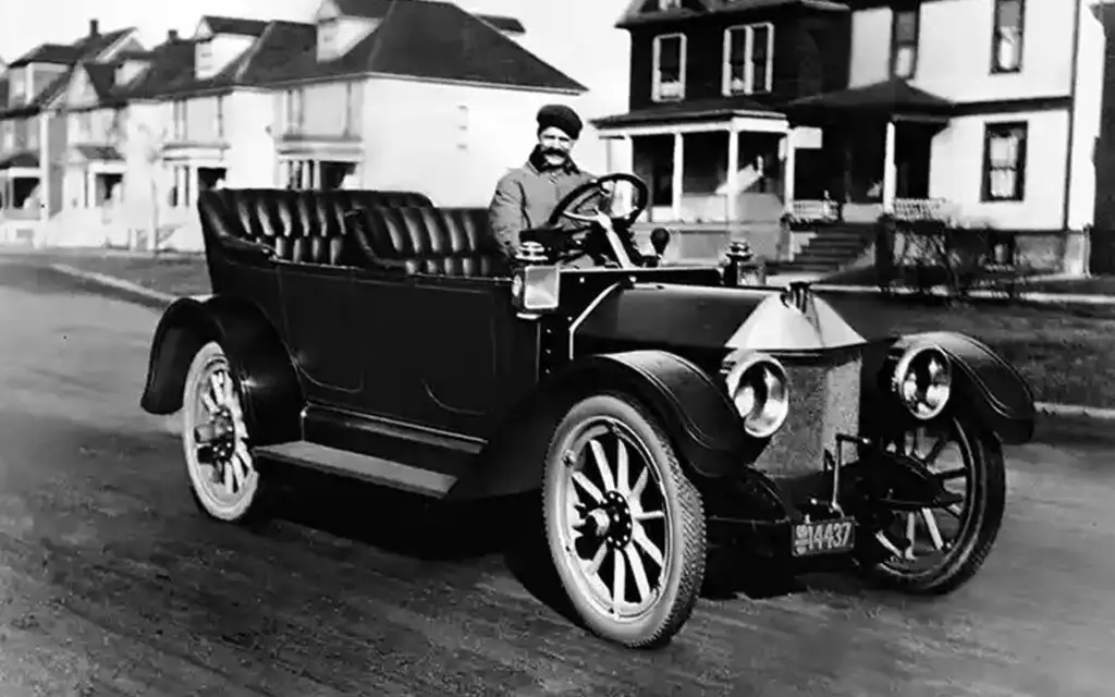 First Cars of Leading Automobile Brands _ Body 11 (20-7-22) - Engine Stories Vintage car from early 1900s with driver on a suburban street, showcasing historic automotive design and architecture.
