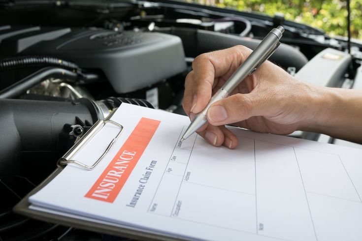 Person filling out an insurance claim form on a clipboard beside a car engine.