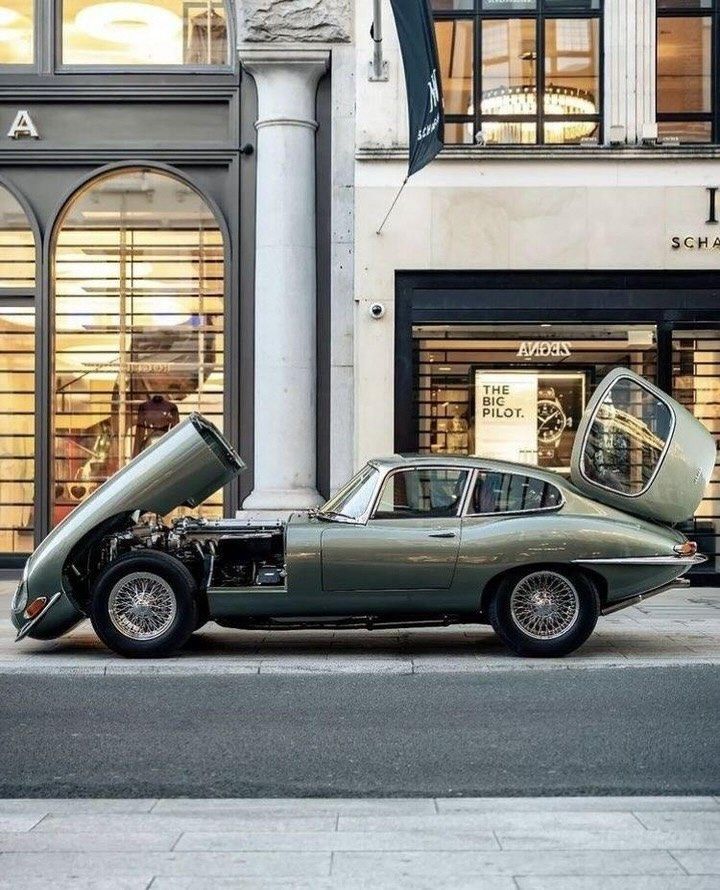 Vintage green sports car with open hood and trunk parked on a city street near a modern store.