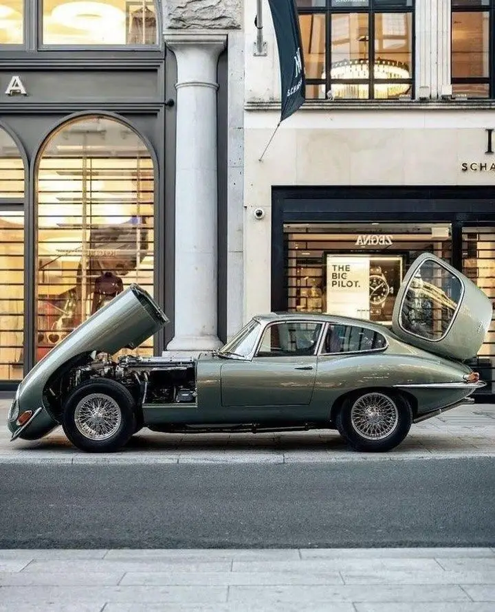 Jaguar Type E 4_2 V8 1961 - Engine Stories Vintage green sports car with open hood and trunk parked on a city street near a modern store.
