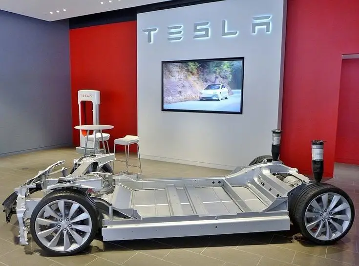 More On US LFP Battery Production Deal With Mystery Automaker - Engine Stories Tesla EV chassis display in a showroom with brand logo and a screen showing a vehicle in motion.
