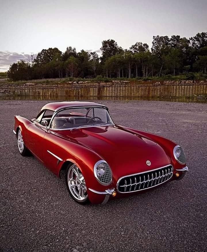 Red classic car parked on a gravel lot, surrounded by trees at twilight, showcasing vintage automotive design.