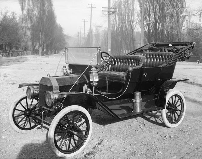 Vintage black and white photo of an old-fashioned car with a convertible top, parked on a dirt road lined with trees.