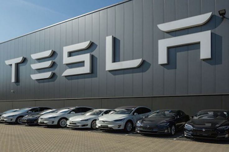 Tesla cars parked outside a large Tesla factory building with logo visible, showcasing modern electric vehicles in a sleek lineup.