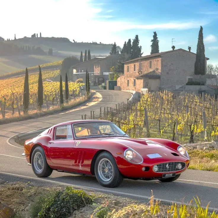 The 1962 Ferrari 250 GTO is widely regarded as one of the most legendary and sought-after cars_ - Engine Stories Classic red car on a scenic countryside road with vineyards and rustic houses in the background under a clear blue sky.