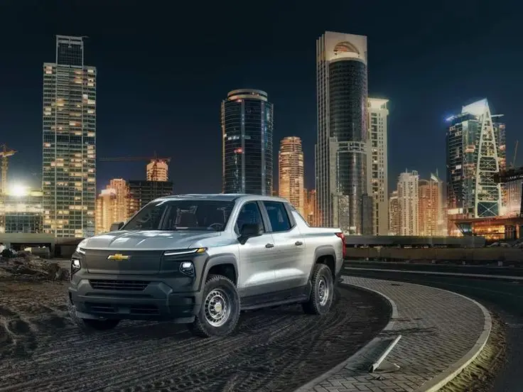 This Is The 2024 Chevrolet Silverado EV Pickup - Engine Stories Silver pickup truck parked on a city street at night, with illuminated skyscrapers in the background.