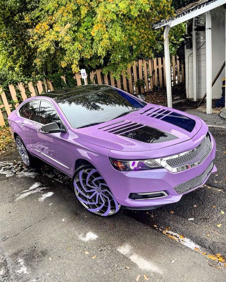 Purple custom car with stylish rims and hood vents, parked outdoors on a wet surface with green foliage in the background.