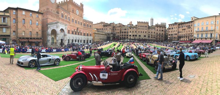 Vintage cars display in historic European square, people admire classic automobiles, medieval architecture in background.