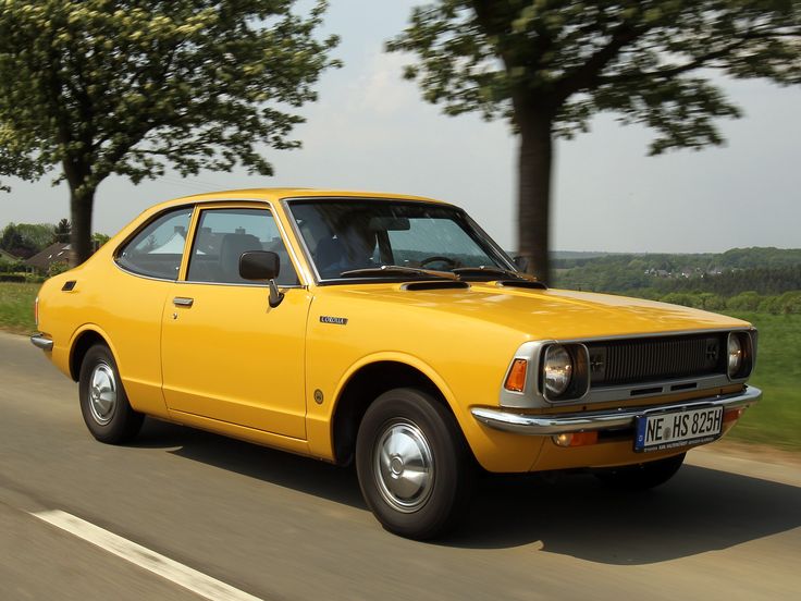 Yellow vintage car driving on a scenic road with trees and horizon in the background, showcasing classic automobile design.