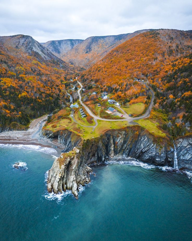 Aerial view of a coastal village beside rugged cliffs and vibrant autumn forests, overlooking a calm ocean.