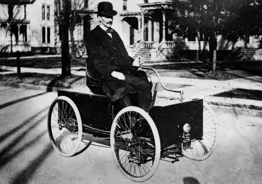 1896-ford-quadricycle-1 - Engine Stories Man in a bowler hat driving a vintage early automobile on a suburban street, showcasing early 1900s transportation history.