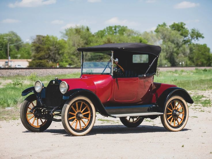 Red vintage car with a black roof parked on a gravel path, surrounded by green trees and a bright blue sky.