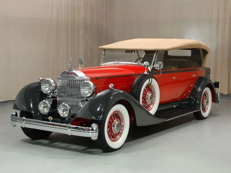 1934 Packard 12 - Engine Stories Vintage red and black classic car with whitewall tires and tan convertible top in a showroom setting.