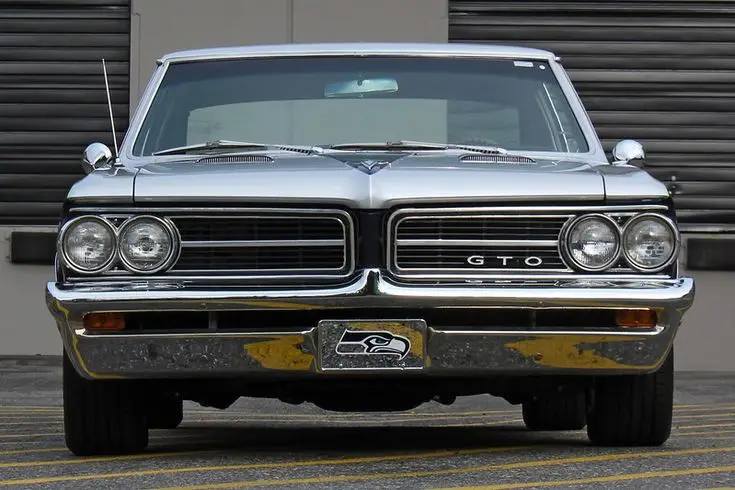 1964 Pontiac GTO - Engine Stories Classic silver Pontiac GTO front view with badge, showcasing iconic design and vintage appeal in a parking lot setting.