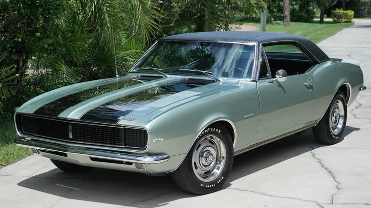 1967 Chevrolet Camaro Z28 - Engine Stories Classic 1967 green Chevrolet Camaro with black racing stripes, parked on a driveway surrounded by lush greenery.