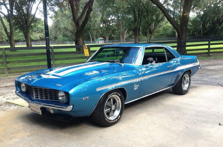 1969 Yenko Camaro - Engine Stories Blue vintage muscle car with white racing stripes, parked on a driveway surrounded by trees and a wooden fence. Classic automotive design.