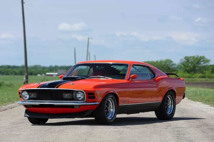 1970 Ford Mustang Mach 1 - Engine Stories Vintage orange muscle car on a rural road under blue skies; classic automotive design with black racing stripes and chrome wheels.