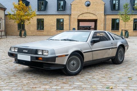 Silver classic DeLorean sports car parked on cobblestone in front of a brick building with modern features.