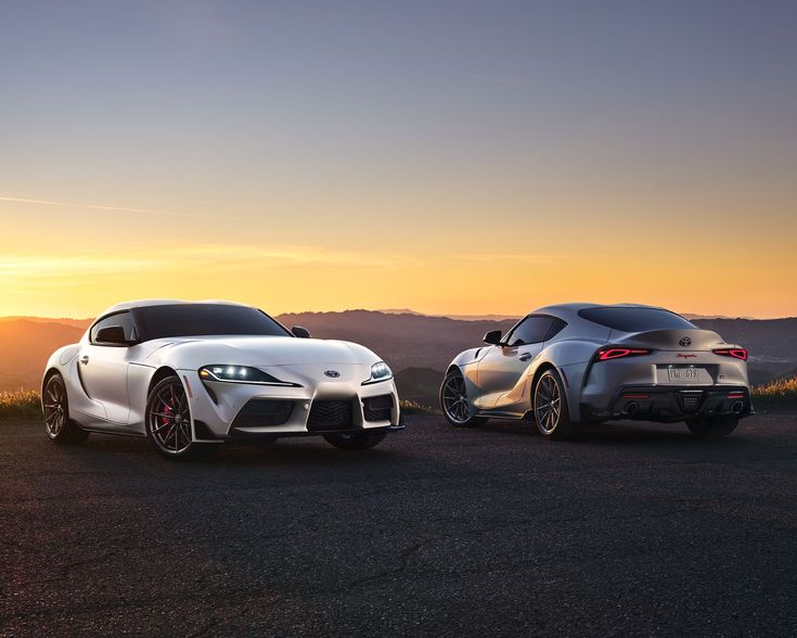 Two sleek white sports cars parked on a scenic overlook at sunset, highlighting their modern design and elegance.