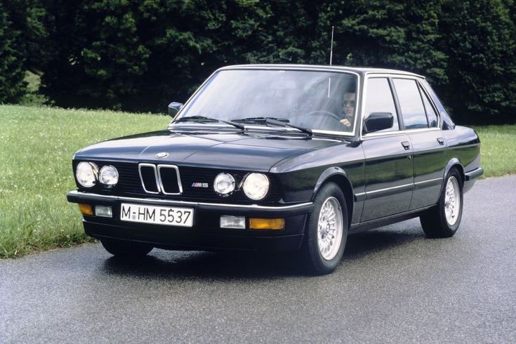 Classic black BMW E28 M5 driving on a scenic road, showcasing vintage design and style in a lush green setting.