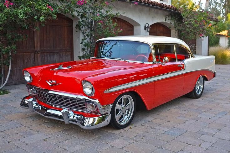 56 Chevy Bel Air - Engine Stories Vintage red and white classic car parked on a cobblestone driveway in front of a rustic garage with flowers.