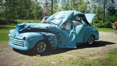Unique blue custom car with open doors and hood, styled like waves, parked outdoors on a sunny day.