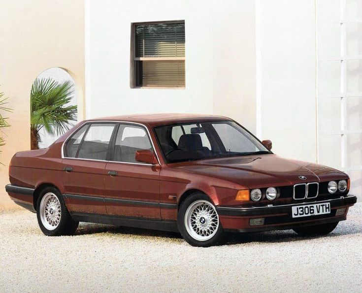 Classic red BMW E32 parked in front of a modern white building with lush green plants, showcasing vintage car design.