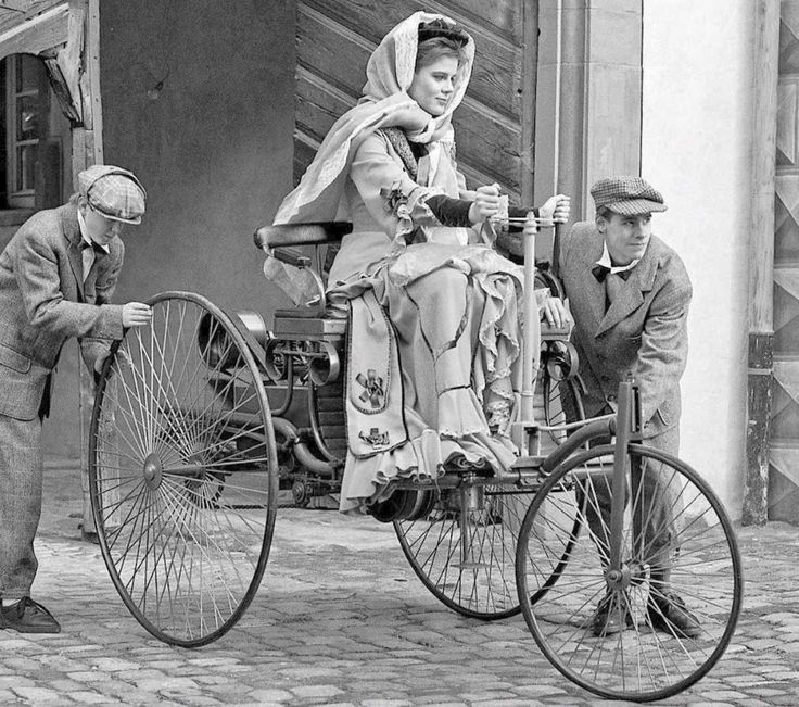 Bertha Benz mujer pionera en el mundo del automóvil - Engine Stories A woman in vintage attire rides an early motorized tricycle, assisted by two men, showcasing historic transportation.