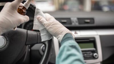 Gloved hands disinfecting a car steering wheel with spray and cloth, ensuring vehicle cleanliness and hygiene.