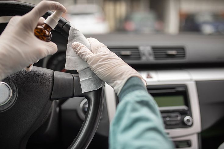 Gloved hands disinfecting a car steering wheel with spray and cloth, ensuring vehicle cleanliness and hygiene.