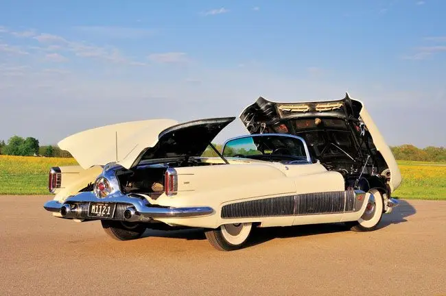 Classic white vintage car with open trunk and hood, parked in scenic countryside under a clear blue sky.