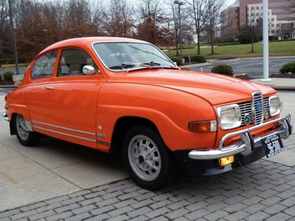 Vintage orange Saab 96 car parked on a paved road, set against a backdrop of trees and buildings.