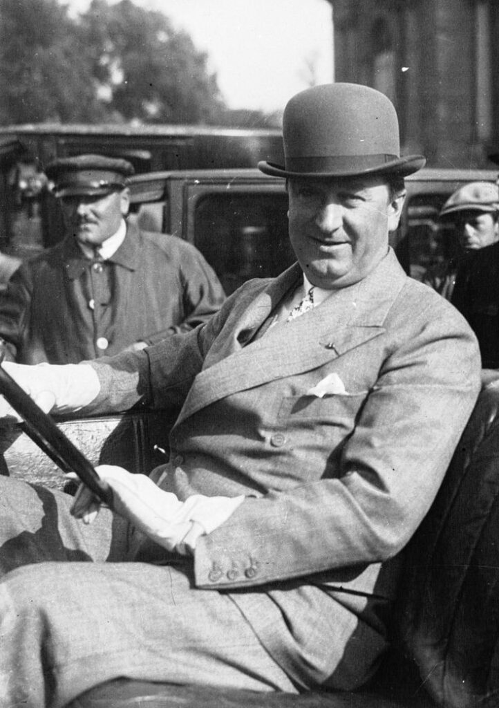 Man in bowler hat and suit smiles while sitting in a vintage car, surrounded by people in early 20th-century attire.