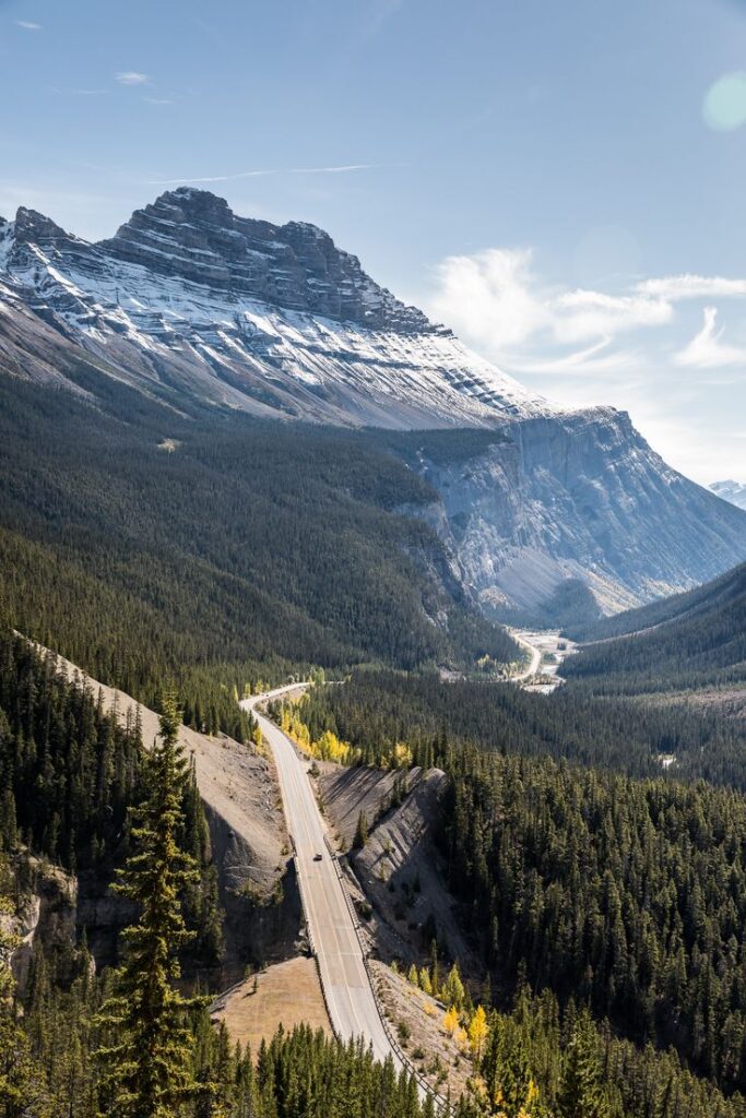 Scenic mountain road with snow-capped peaks and dense forest below a blue sky in Banff National Park, Canada.