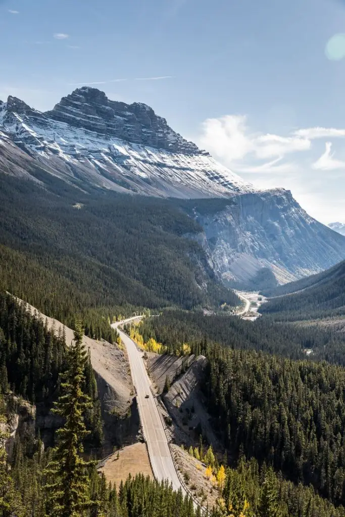 Experience the Icefields Parkway_ A Scenic Drive of a Lifetime - Engine Stories Scenic mountain road with snow-capped peaks and dense forest below a blue sky in Banff National Park, Canada.