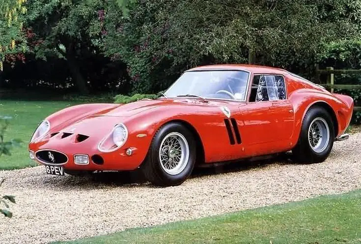 Ferrari 250 GTO 1962 - Engine Stories Red vintage sports car parked on a gravel path surrounded by lush greenery, showcasing classic design and elegance.