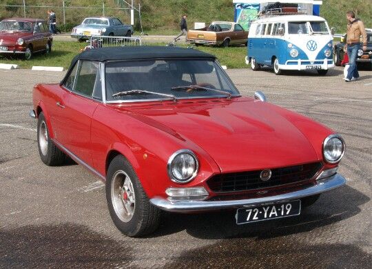 Fiat 124 Spider 1971 - Engine Stories Classic red convertible car parked outdoors with vintage vehicles in the background, showcasing timeless automotive style.