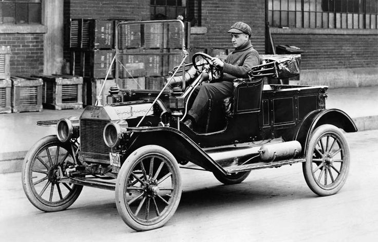 Ford Model T - Engine Stories Vintage black-and-white photo of a man driving a classic Model T Ford car on a city street, showcasing early 20th-century automobiles.