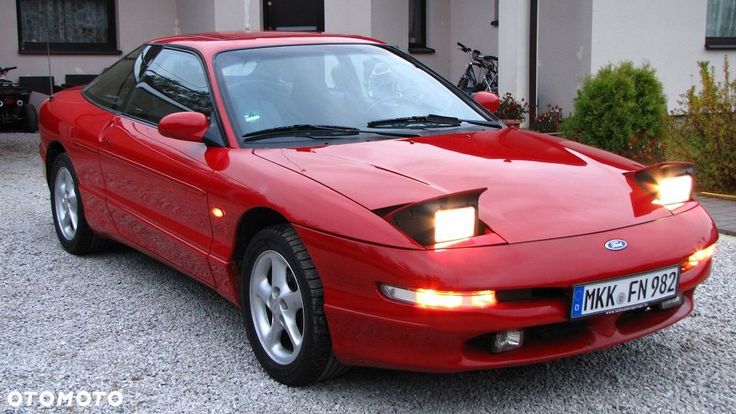 Ford Probe - Engine Stories Red classic sports car with pop-up headlights, parked on a gravel driveway in front of a house.
