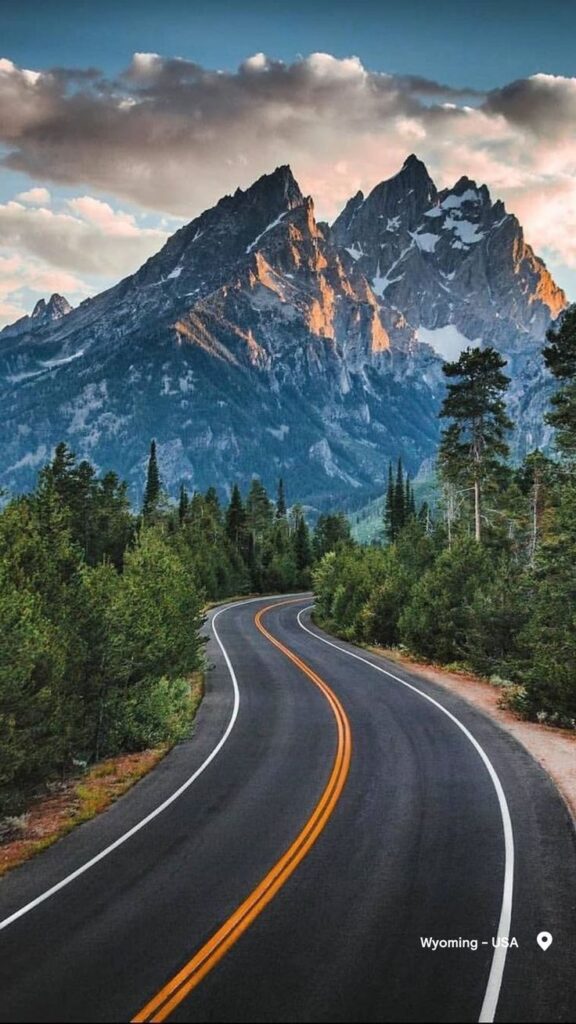 Scenic winding road leading to majestic mountains in Wyoming, USA, framed by lush green trees under a cloudy sky.