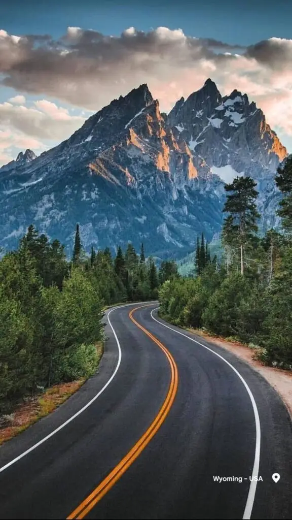 Grand Teton National Park - Engine Stories Scenic winding road leading to majestic mountains in Wyoming, USA, framed by lush green trees under a cloudy sky.