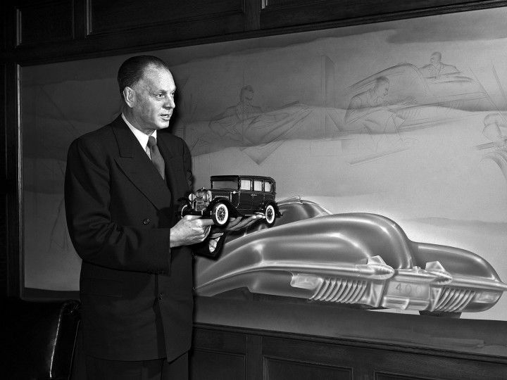 Man holding vintage model car in front of futuristic car design sketch, showcasing automotive innovation and history in black and white.