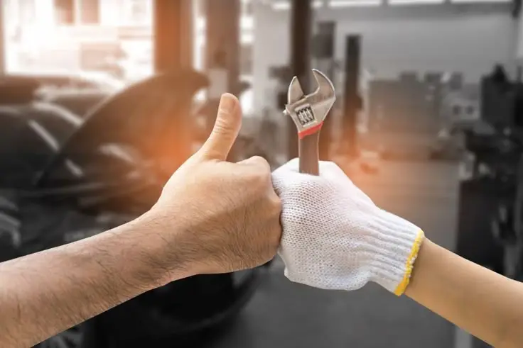 Thumbs up and wrench handshake in a workshop, symbolizing teamwork and cooperation in automotive repair, with warm lighting.