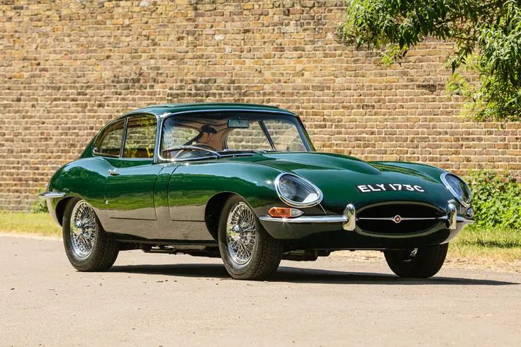 Classic green sports car parked by brick wall, showcasing vintage design and chrome wheels in sunny outdoor scene.