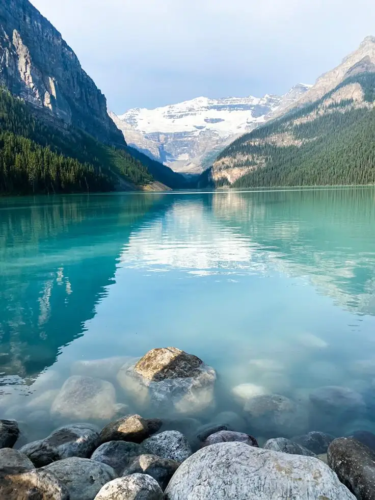 Lake Louise Canada - Engine Stories Serene mountain lake with clear turquoise water, rocky shoreline, and snow-capped peaks in the background on a clear day.