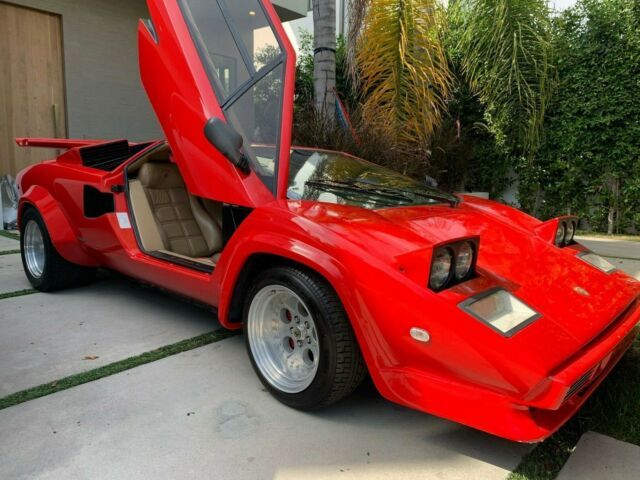 Red sports car with iconic scissor doors open, parked in a driveway surrounded by lush greenery.